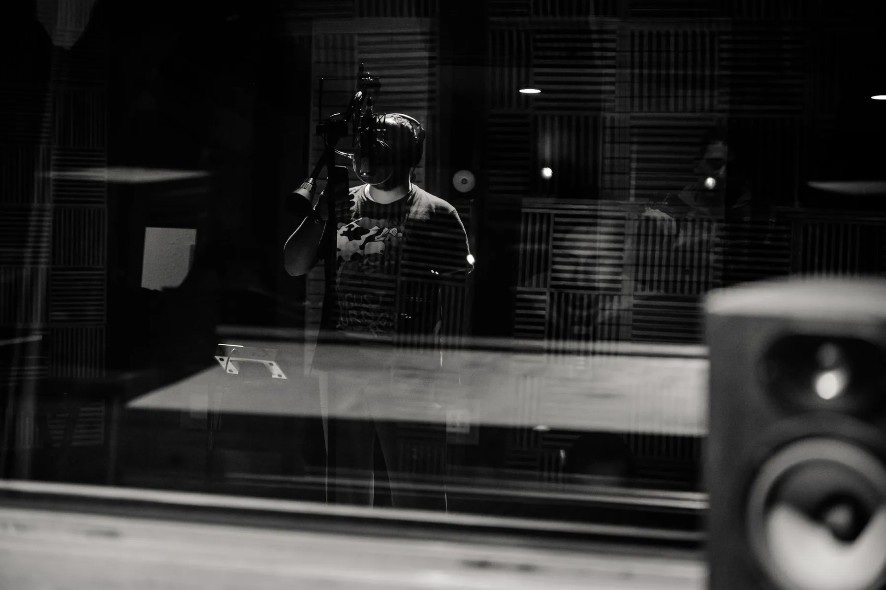 A man singing in a vocal booth, seen through the control room glass, in black and white.