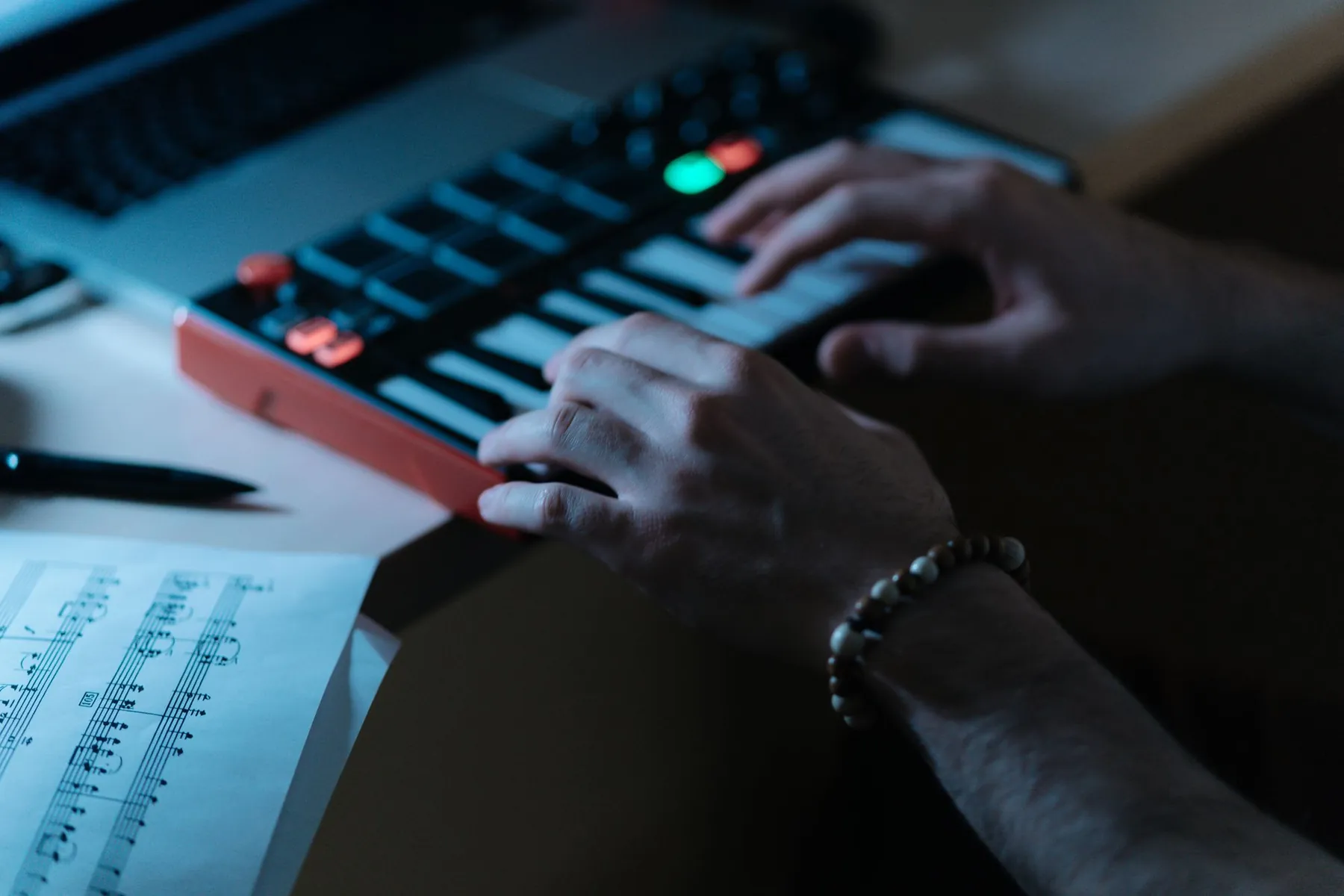 Hands playing a small MIDI keyboard beside a laptop at night.