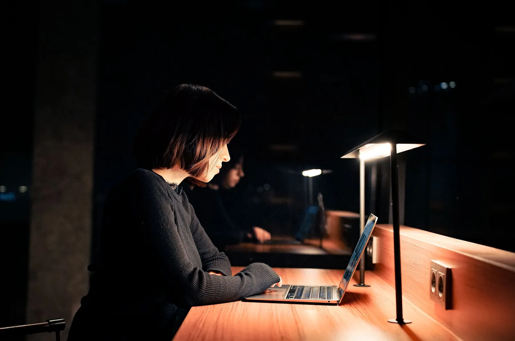 A woman working on a laptop under a desk lamp at night.