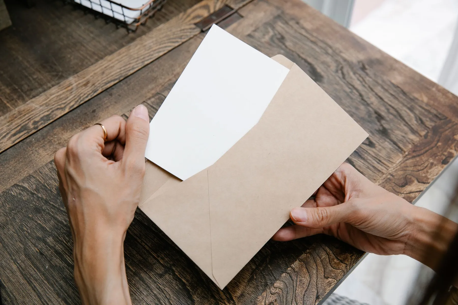 Hands holding a card in a kraft envelope on a wooden desk.