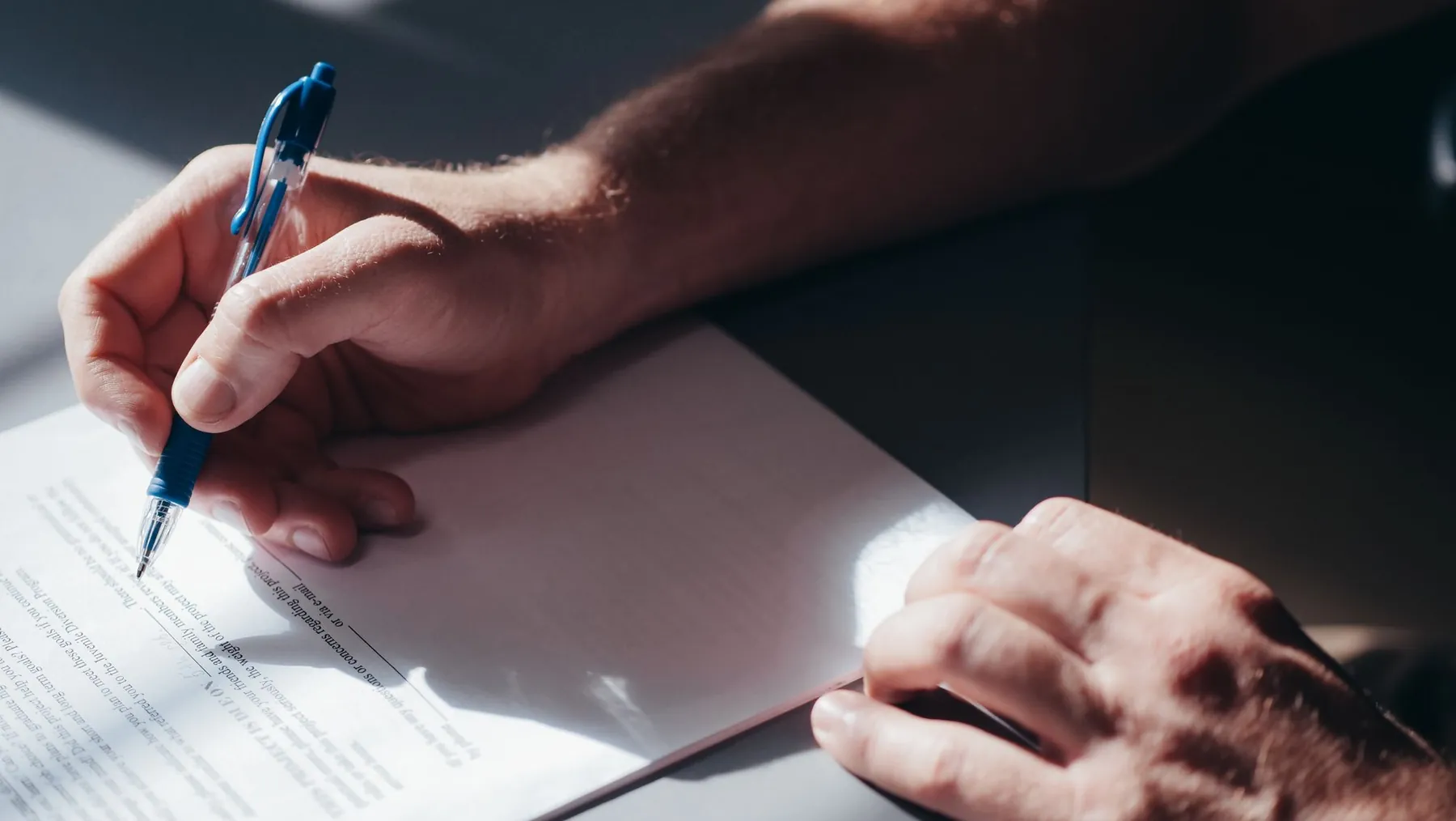 Hands signing a contract with a blue pen in sunlight.